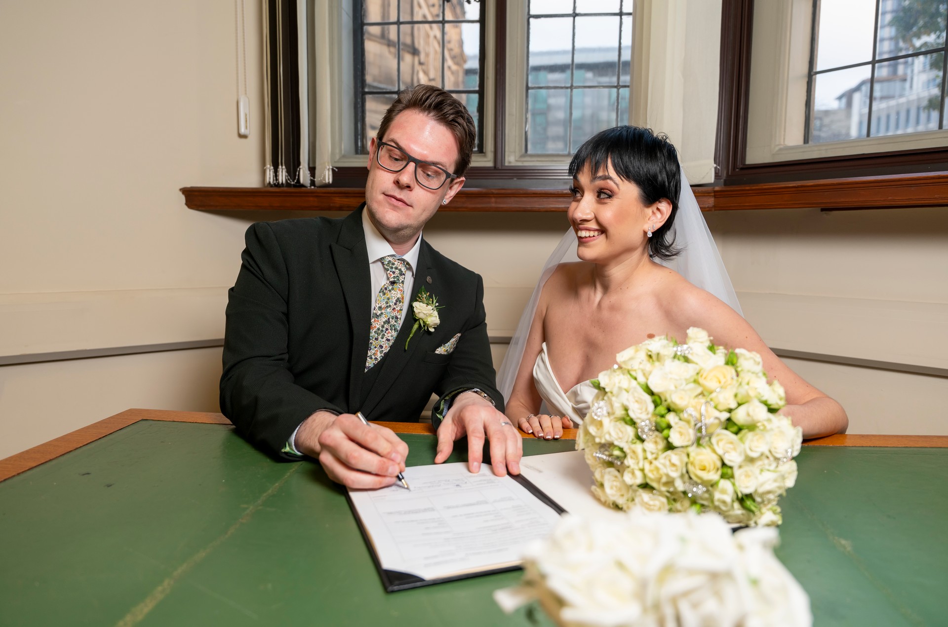 groom in black suit and tie signs his wedding certificate at indoor ceremony, smiling bride dressed in white gown holding bouquet of flowers